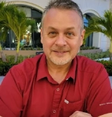 A man in red shirt sitting outside with palm trees.